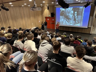 Fullsatt auditorium under et skoleforedrag, lerret med bilde foran.